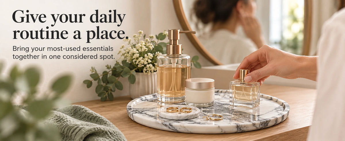 Hand reaching for perfume on a grey marble vanity tray holding a soap dispenser, skincare jar, and jewelry.