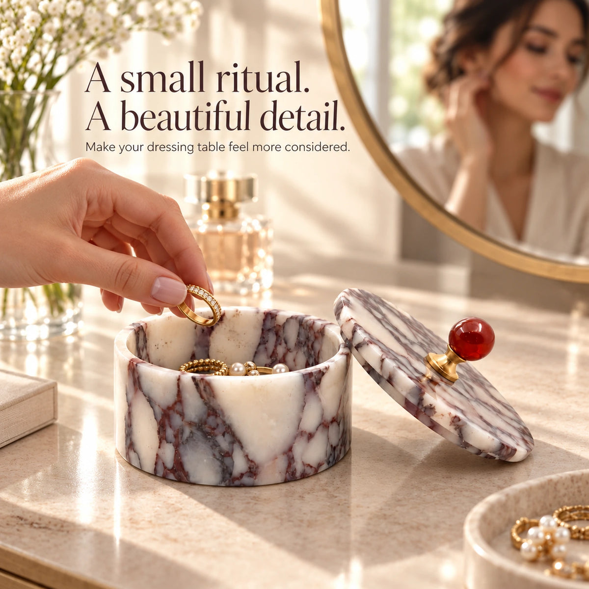 Hand placing rings into an open purple marble jewelry box on a vanity beside perfume, flowers and a round mirror.