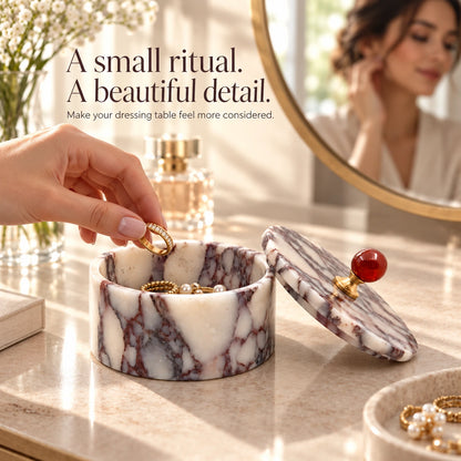 Hand placing rings into an open purple marble jewelry box on a vanity beside perfume, flowers and a round mirror.