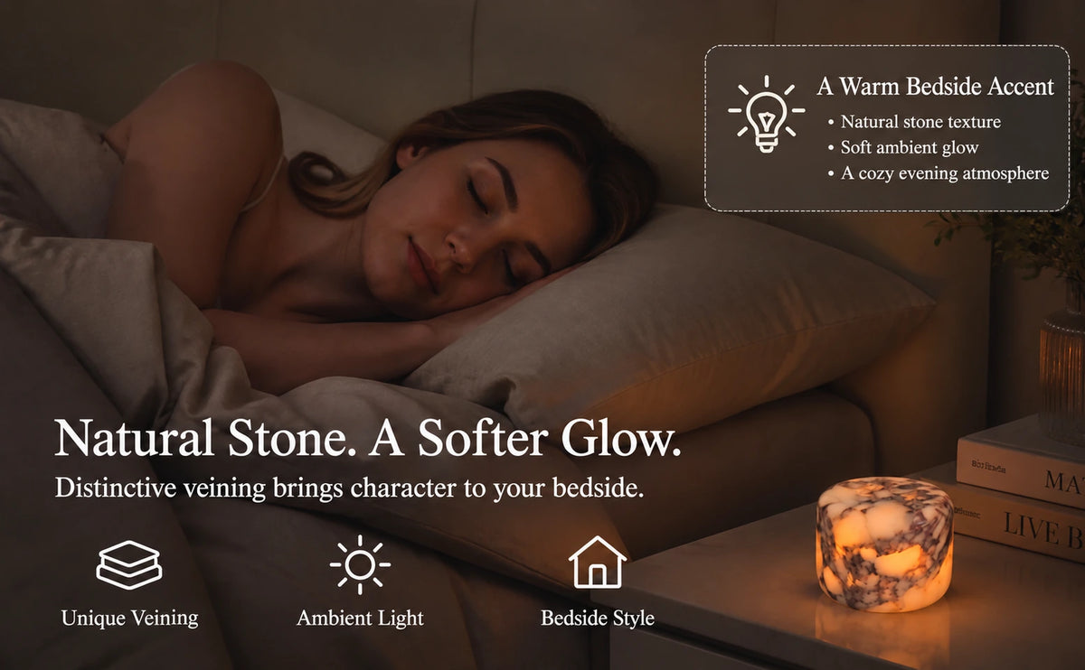 Natural stone lamp casting a warm amber glow on a bedside table beside books and a sleeping woman.