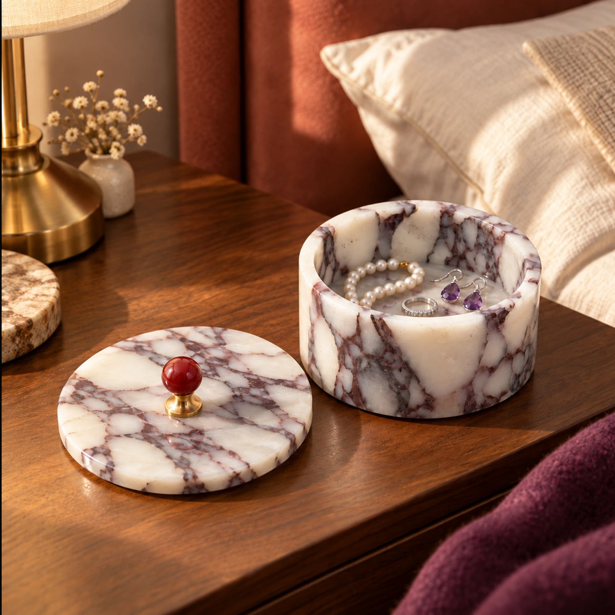 Open purple marble jewelry box holding a pearl bracelet, ring and earrings on a wooden bedside table, with its red-knob lid beside it.